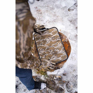 Person wearing Mobile Warming camouflage gloves holding a camouflage heated seat pad on a tree stump in the snow