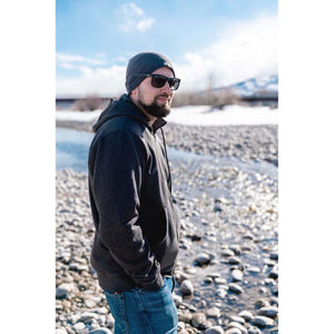 Man wearing a Mobile Warming dark grey hooded sweatshirt and beanie standing by a rocky river