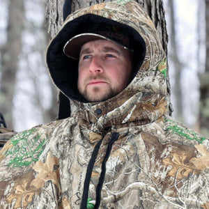 Man wearing Realtree camouflage hooded hunting suit and Heater Body Suit cap in a forest