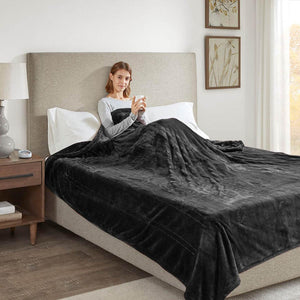 Person sitting on a bed covered by a dark grey plush electric blanket holding a mug