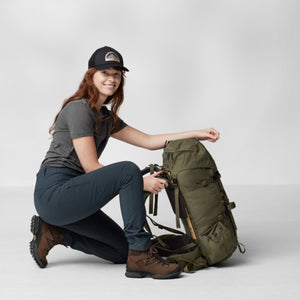 Woman wearing FjallRaven cap and hiking gear kneeling beside an olive green backpack