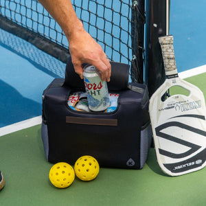 Hand reaching into a black cooler bag with Coors Light cans next to a Selkirk pickleball paddle and two yellow balls