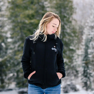Woman wearing a black Go! Heat zip up hoodie with blue jeans in a snowy forest