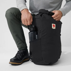 Person placing a black water bottle into the side pocket of a black Fjallraven backpack
