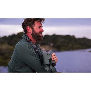 Man with beard wearing green long sleeve shirt holding binoculars by a river