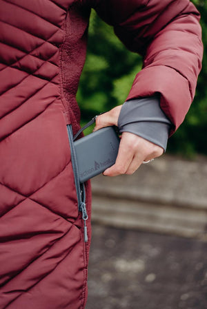 Hand placing an ActionHeat power bank into the zippered pocket of a quilted maroon jacket