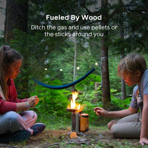 Two children sitting on the ground by a small portable wood burning camping stove in a forest