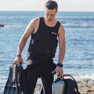 Man wearing a Venture Heat vest and wetsuit holding scuba diving fins and a tank by the ocean